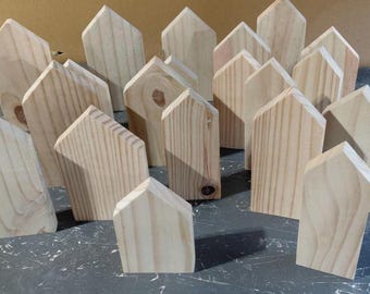 May include: A collection of unfinished light-colored wooden house-shaped cutouts. Each piece features a pointed roof and a rectangular base, displaying natural wood grain patterns and knots. The set includes multiple pieces of varying heights.