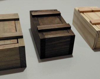 May include: Three rectangular wooden boxes designed to resemble miniature shipping crates. Each box features a sliding lid with two horizontal wooden slats across the top. The boxes are finished in three distinct wood tones: light gray, dark brown, and natural light tan.