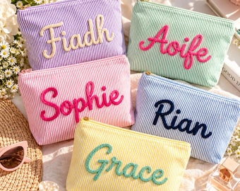 Op de afbeelding: Vijf rechthoekige etuis met rits van gestreepte stof in paars, groen, roze, blauw en geel. Elk etui is voorzien van een in cursief geborduurde naam: Fiadh, Aoife, Sophie, Rian en Grace, uitgevoerd in contrasterende garenkleuren.