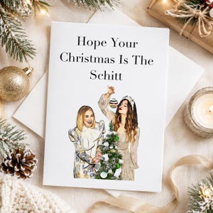Könnte beinhalten: Eine weiße Grußkarte mit dem schwarzen Text Hope Your Christmas Is The Schitt über einer farbigen Illustration von zwei Frauen, die einen kleinen Weihnachtsbaum mit Kugeln schmücken, eine trägt ein gemustertes Oberteil und die andere einen hellen Pullover.