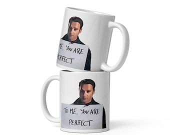 May include: Two white ceramic mugs stacked vertically, each featuring a printed illustration of a man holding a white sign with the handwritten black text, "TO ME, YOU ARE PERFECT." The mugs have a standard cylindrical shape and a side handle.