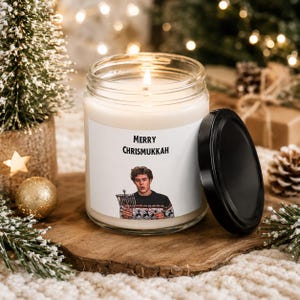 May include: A clear glass candle jar with a white wax fill and a black lid. The label features the text Merry Chrismukkah above an illustration of a person wearing a patterned sweater and holding a silver menorah. The candle has a lit wick.