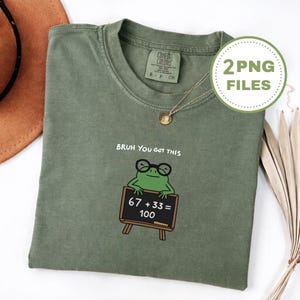 May include: Olive green t-shirt featuring a frog with glasses and a chalkboard displaying "67 + 33 = 100". The phrase "BRUH YOU GOT THIS" is above the frog. A gold necklace is also visible.