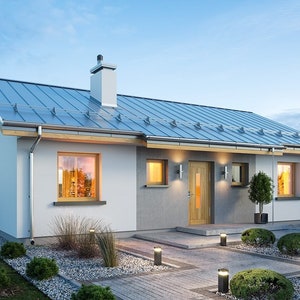 May include: A single-storey white house with a grey metal roof and a wooden front door. The house has a small front garden with landscaping and a gravel driveway.