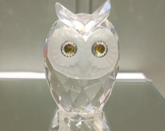 May include: A clear crystal owl figurine with yellow eyes, displayed on a mirrored surface atop a white box. The box is labeled "VDF ANTIQUES & COLLECTIBLES". The owl has a faceted design, reflecting light.