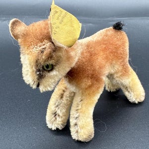 以下が含まれることがあります： ヴィンテージのぬいぐるみ、鹿の形。おもちゃは主に茶色で、ベージュの脚と黒い尻尾があります。耳には黄色のタグが付いています。柔らかく、豪華な素材で作られています。