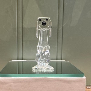 Puede incluir: Una figura de perro de cristal transparente con ojos y nariz negros, de pie sobre una superficie de vidrio. La figura se exhibe en una caja con el texto "VDF ANTIQUES & COLLECTIBLES". El fondo es una pared verde claro.