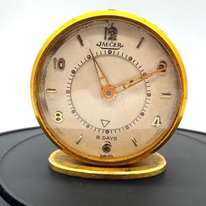 May include: A round gold-toned Jaeger desk clock with a cream-colored face, featuring the text JAEGER, 8 DAYS, and SWISS. The clock has gold-toned hands, Arabic numerals at the 12, 10, 8, 4, and 2 positions, and a small oval base stand.