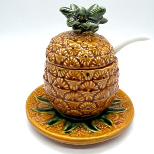May include: A ceramic pineapple-shaped sugar bowl with a lid and a small spoon. The bowl is brown with a textured surface, and the lid is topped with green leaves. It sits on a matching saucer with green leaf accents.
