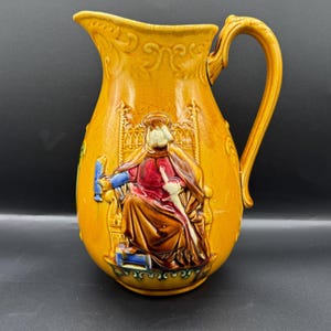 Peut inclure: Pichet en céramique jaune moutarde avec une anse. Le pichet présente un relief en relief d'une figure assise en robe, tenant un bâton et un objet bleu. Le pichet a un bec et un bord décoratif.