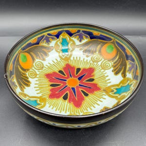 Puede incluir: Un cuenco de cerámica pequeño y redondo con un diseño floral pintado a mano. El cuenco presenta una flor roja y naranja en el centro, rodeada de motivos de plumas de pavo real en tonos marrones, verdes y azules. El borde es marrón oscuro.