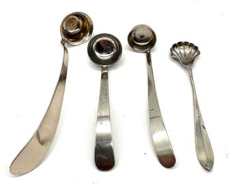 Puede incluir: Cuatro cucharas de servir antiguas de plata. Las cucharas tienen diferentes formas de cuenco, incluyendo diseños redondos y festoneados. Los mangos son delgados y curvos. Las cucharas están dispuestas sobre una superficie blanca.