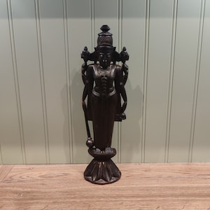 Può includere: Una statua in legno marrone scuro di una divinità indù con quattro braccia, in piedi su un piedistallo con una base scolpita. La statua è riccamente dettagliata e ha una corona sulla testa.