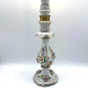 May include: A white porcelain table lamp featuring a bulbous base decorated with raised, hand-painted floral motifs in pink, blue, and green with gold trim. The lamp has a fluted stem, a scalloped base, and a white power cord with a toggle switch.