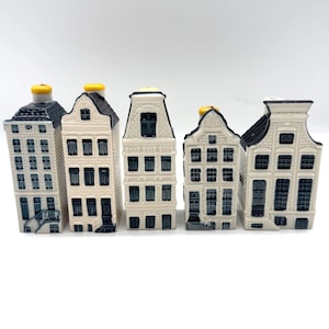 Puede incluir: Una colección de cinco figuritas de casas de cerámica. Cada casa en miniatura presenta un diseño único con diferentes detalles de ventanas y tejados. Las casas son principalmente blancas con detalles en negro y azul, y algunas tienen chimeneas amarillas.