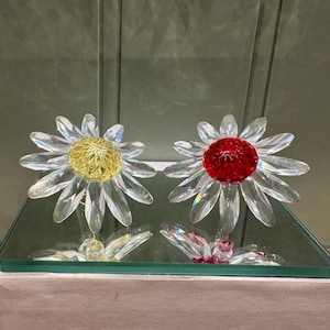 以下が含まれることがあります： 透明な花びらと黄色と赤の中心を持つ2つのガラス製デイジー彫刻。 花は透明なガラスベースに取り付けられ、白い箱の上に置かれています。 デイジーは装飾品です。