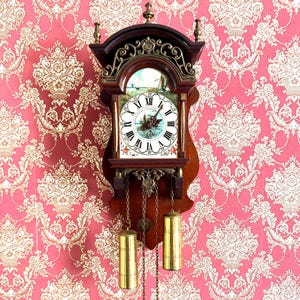 Könnte beinhalten: Eine Vintage-Wanduhr aus Holz mit dekorativem Zifferblatt und Pendel. Die Uhr hat eine dunkelbraune Oberfläche mit goldenen Akzenten und zwei messingfarbenen Gewichten. Sie hängt vor einem rosa Tapetenhintergrund mit floralem Muster.