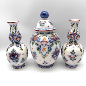 May include: A set of three white ceramic vessels featuring hand-painted floral patterns in red, blue, and green. The collection includes two hourglass-shaped vases and one central lidded jar topped with a round blue finial, all with ornate detailing.