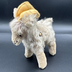 Può includere: Un giocattolo vintage a forma di montone di peluche con pelliccia marrone e un cappello di feltro giallo. Il montone ha un bottone sul lato e sta in piedi sulle quattro zampe. Il giocattolo è realizzato in materiale morbido e felpato.