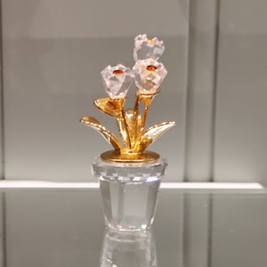 Può includere: Un'arrangiamento di fiori di cristallo con tre fiori bianchi e foglie dorate in un vaso di cristallo trasparente.