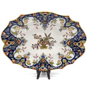 Op de afbeelding: Een ovale keramische schaal met een geschulpte rand en gouden afwerking. Het midden toont een illustratie van een bloemenmand op een witte achtergrond, omringd door een brede rand met donkerblauwe, groene en oranje bloem- en krulpatronen.