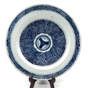Puede incluir: Un plato de cerámica blanco y azul con un diseño floral detallado. El plato presenta un motivo floral central y un borde estampado. El plato se exhibe sobre un soporte de madera, mostrando sus intrincados detalles y artesanía.