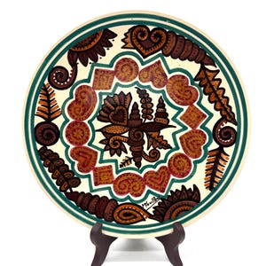 Könnte beinhalten: Ein dekorativer Keramikteller mit cremefarbenem Hintergrund und grünem Rand. Der Teller zeigt ein symmetrisches Design aus braunen und roten Formen, darunter Herzen, Quadrate und florale Elemente. Der Teller wird auf einem dunkelbraunen Holzständer präsentiert.
