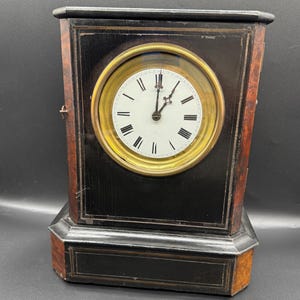 May include: An antique black mantel clock with a gold-coloured clock face and Roman numerals. The clock has a rectangular shape with a wooden base and sides. The clock face is white with black hands.