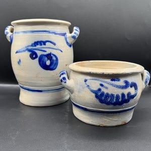 May include: Two antique stoneware pottery pieces. The larger piece is a tall, cylindrical jar with two handles and blue floral designs. The smaller piece is a round bowl with two handles and similar blue designs. Both pieces have a light beige base color.