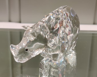May include: A clear, faceted crystal figurine of a rhinoceros. The animal is depicted in a standing position with a prominent horn on its snout and a small, dark, circular eye detail. The crystal surface features multiple geometric cuts.