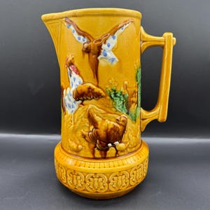 Peut inclure: Pichet en céramique jaune moutarde avec un motif en relief détaillé. Le pichet comporte une anse et une base avec un motif en relief. Le relief représente un coq, un oiseau en vol et d'autres animaux de la ferme. Le pichet est un objet décoratif.