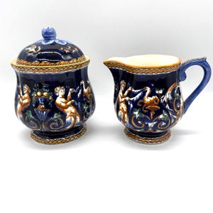 May include: A dark blue ceramic sugar bowl with a lid and a matching creamer. Both pieces feature a decorative design with figures, foliage, and gold accents. The creamer has a blue handle and a light-colored rim.