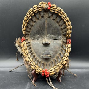 May include: A dark wooden mask with a serene expression, decorated with a ring of seashells and red beads. The mask is mounted on a small wooden stand, highlighting its intricate details and cultural significance.