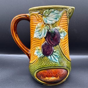 Peut inclure: Pichet en céramique avec une anse, orné d'un motif de prunes et de feuilles. Le pichet a une anse marron, un corps jaune et vert, et un motif de fruits de couleur prune foncée. Le pichet mesure environ 20 cm de haut.