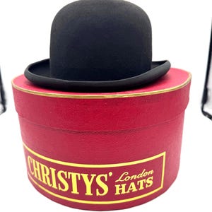 May include: A black bowler hat with a rounded crown and a curved brim, resting on a cylindrical red hat box. The box features a gold-bordered label with the text CHRISTYS' London HATS printed in yellow lettering against a red background.