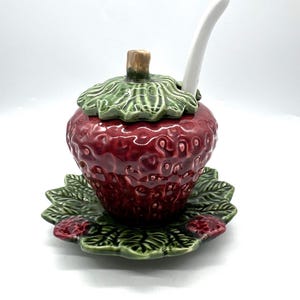 Puede incluir: Un azucarero de cerámica con forma de fresa, con tapa y cuchara a juego. El cuenco es de color rojo intenso con una parte superior verde en forma de hoja y se asienta sobre un platillo verde en forma de hoja. La cuchara es blanca.