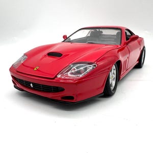 May include: A red Ferrari model car featuring a sleek aerodynamic body, clear headlights, a black front grille with a small Ferrari emblem, a hood scoop, and silver wheels. The vehicle is captured from a front three-quarter angle, showcasing its glossy finish and detailed exterior design elements on a plain white background.