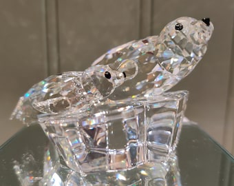 Puede incluir: Una escultura de cristal de dos focas, una adulta y una joven, sobre un iceberg de cristal. Las focas tienen ojos negros y están hechas de cristal facetado, que refleja la luz. La escultura está sobre una superficie espejada.