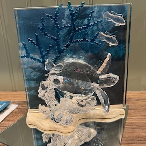 Pode incluir: Uma escultura de cristal de uma tartaruga marinha nadando através de um recife de coral. A tartaruga é feita de cristal transparente e está rodeada de coral de cristal azul. Também há dois peixes de cristal menores nadando no fundo.