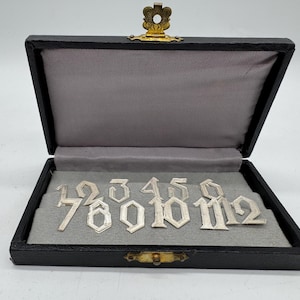 Könnte beinhalten: Eine schwarze Box mit goldenem Verschluss und grauem Innenraum enthält silberne Zahlen von 1 bis 12. Die Zahlen sind in einer dekorativen Schriftart gehalten. Die Box ist geöffnet und zeigt die auf grauem Stoff angeordneten Zahlen.