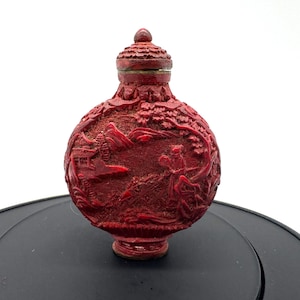 Può includere: Una tabacchiera rotonda rossa con intricati dettagli in rilievo intagliati raffiguranti un paesaggio con una figura, alberi e strutture architettoniche. La bottiglia ha un coperchio rosso abbinato con una superficie testurizzata e un collo stretto.