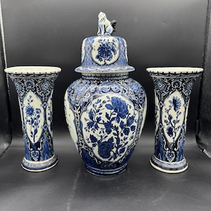 Peut inclure: Ensemble de trois vases en céramique bleus et blancs. Le vase central est un grand pot à couvercle avec des motifs floraux. Deux vases plus petits encadrent le pot. Le couvercle du pot est surmonté d'une figurine de lion.