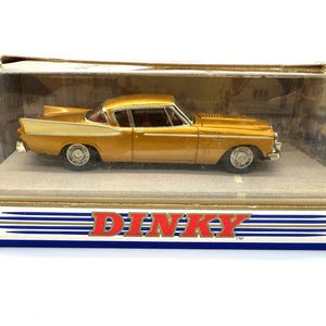 May include: A gold-coloured Dinky die-cast model car with silver trim and tail fins, enclosed in its original clear plastic and cardboard display box. The box features the red Dinky logo on the front base and a circular sticker on the left side.