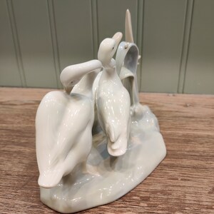 May include: A porcelain figurine of three white ducks standing on a light blue base. The ducks are arranged in a playful pose, with one duck looking back at the other two.