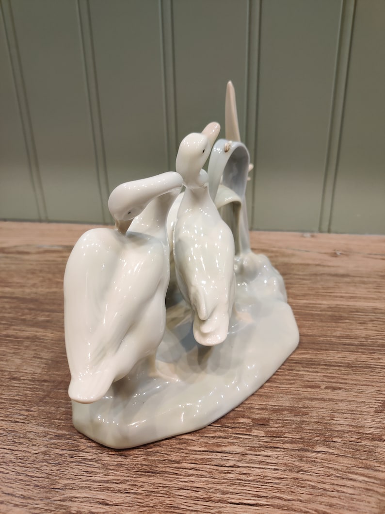 May include: A porcelain figurine of three white ducks standing on a light blue base. The ducks are arranged in a playful pose, with one duck looking back at the other two.