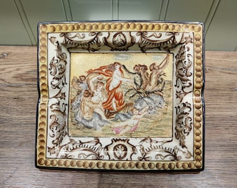 May include: A square ceramic ashtray with a gold and brown border. The center features a colorful illustration of a woman riding a seahorse with other mythical creatures.