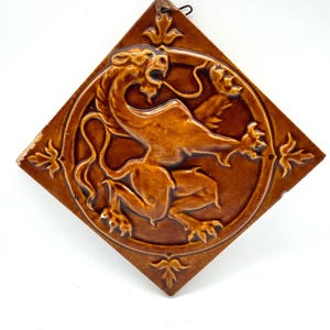 Könnte beinhalten: Eine rautenförmige, braune Keramik-Wandfliese mit einem erhabenen Relief eines Fabelwesens in Form eines Löwen innerhalb einer kreisförmigen Umrandung. Die Fliese hat dekorative Blattmotive an jeder Ecke und eine kleine Metallaufhängung oben.