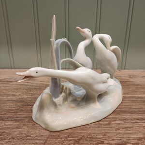 May include: A porcelain figurine of three white geese standing on a base with a light blue and white floral design. The geese are in a playful pose, with one goose looking back at the other two.