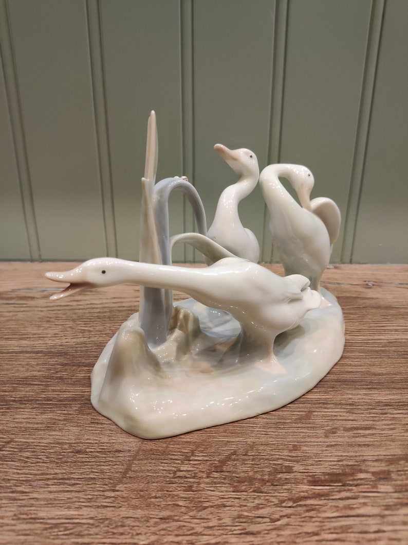 May include: A porcelain figurine of three white geese standing on a base with a light blue and white floral design. The geese are in a playful pose, with one goose looking back at the other two.