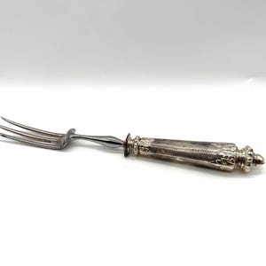 May include: A silver-toned three-tined fork featuring a polished metal head and a textured, ornate handle with intricate detailing. The handle displays a cross-hatched pattern and decorative embossed elements at the base and the end cap.